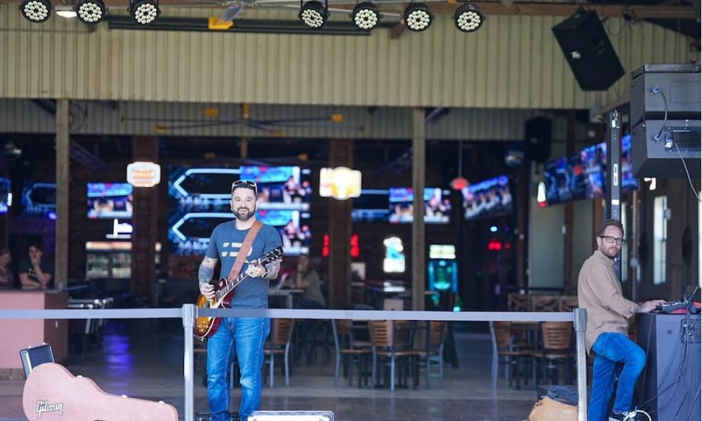 <span class="uk-h4">Musician with guitar performs on outdoor patio stage with professional lighting rig and screens visible in background.</span>