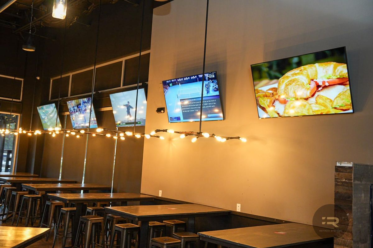 Long wall with multiple TVs above dark tables and hanging string lights in a bar.