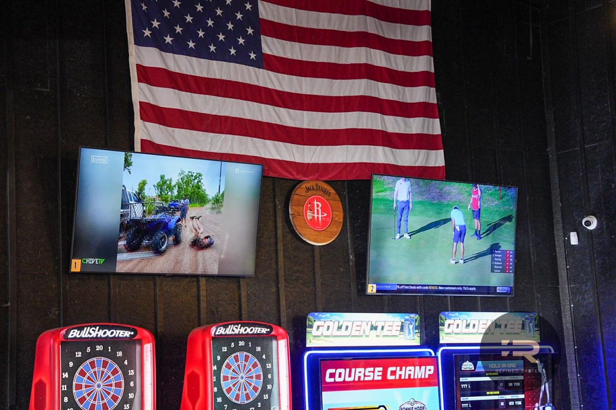 Two TVs playing sports above dart machines and arcade games under a US flag.