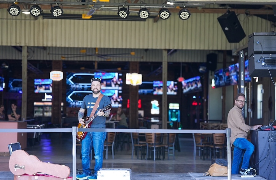 Musician with guitar performs on outdoor patio stage with professional lighting rig and screens visible in background.
