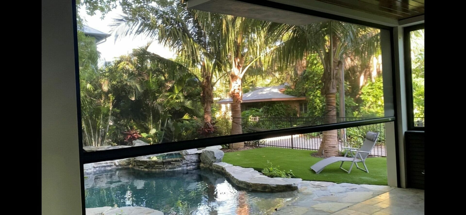patio screen partially lowered over a pool and palm tree backyard with stone patio and lounge chair