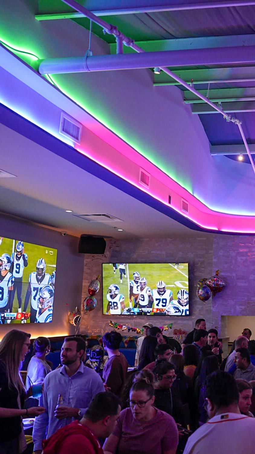 Lively bar with colorful ceiling lighting, wall-mounted TVs, and a crowd of people socializing