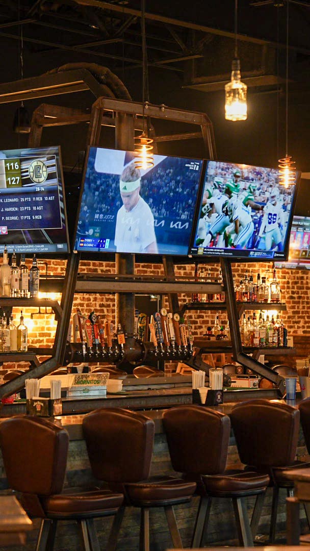 Sports bar with beer taps, brick wall backdrop, and overhead TVs broadcasting games and sports news