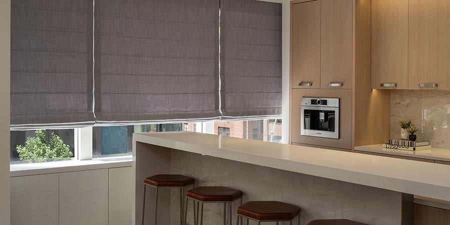 Fabric roman shades lowered over a window behind a modern kitchen island with barstools