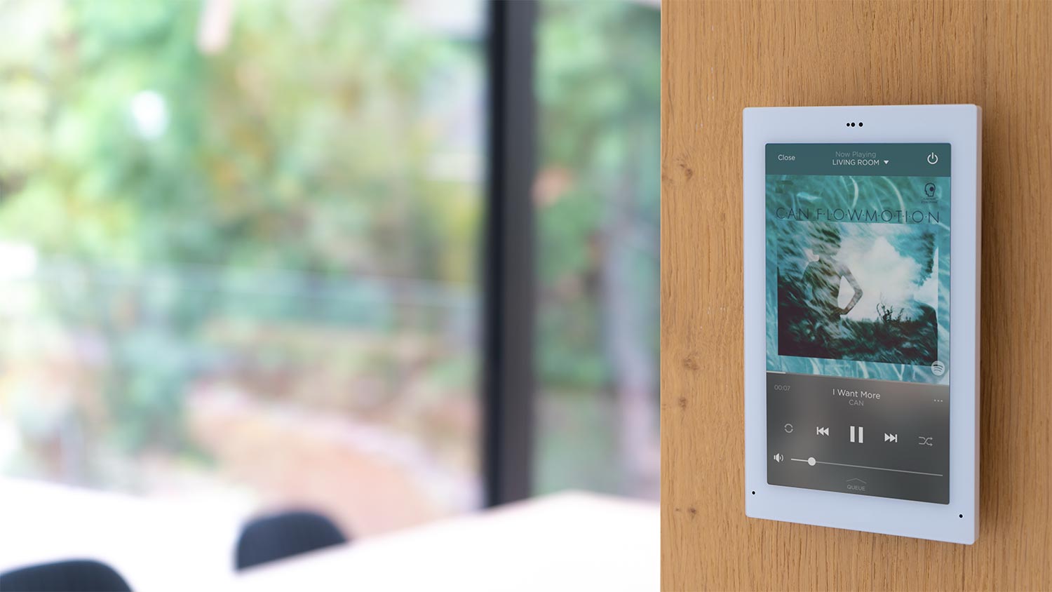 Wall-mounted smart tablet displaying music controls with blurred garden view behind glass