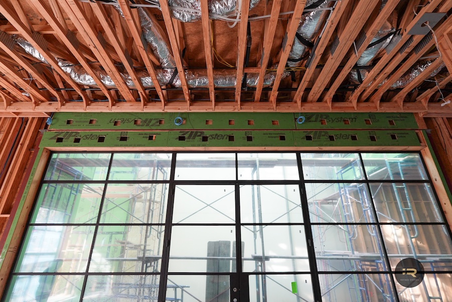 Framed ceiling joists above a large black-framed window wall on a construction site, exposed insulation ducts