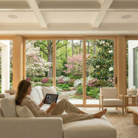 A woman sitting on a white couch, holding a tablet. The room features floor-to-ceiling windows, showing cherry blossom trees outside.