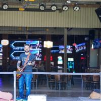 Musician with guitar performs on outdoor patio stage with professional lighting rig and screens visible in background.