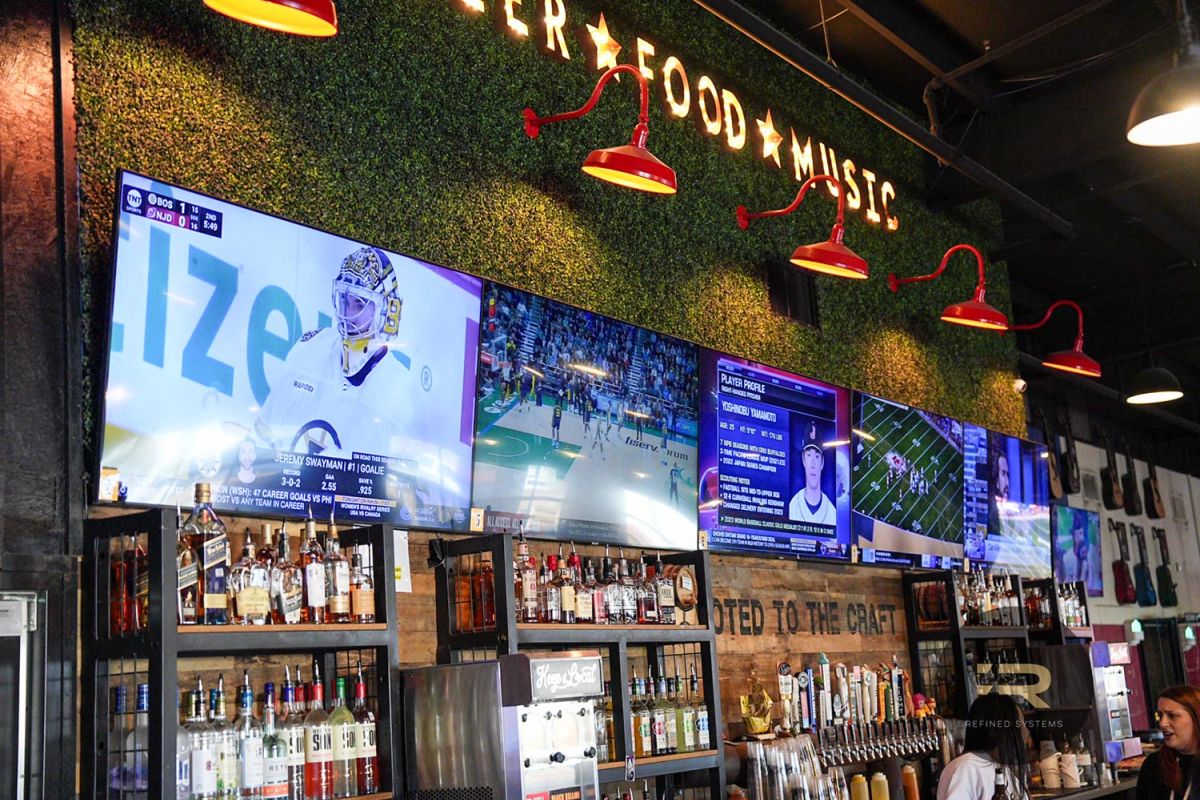 Row of TVs above a bar with liquor bottles and sports showing on the screens.