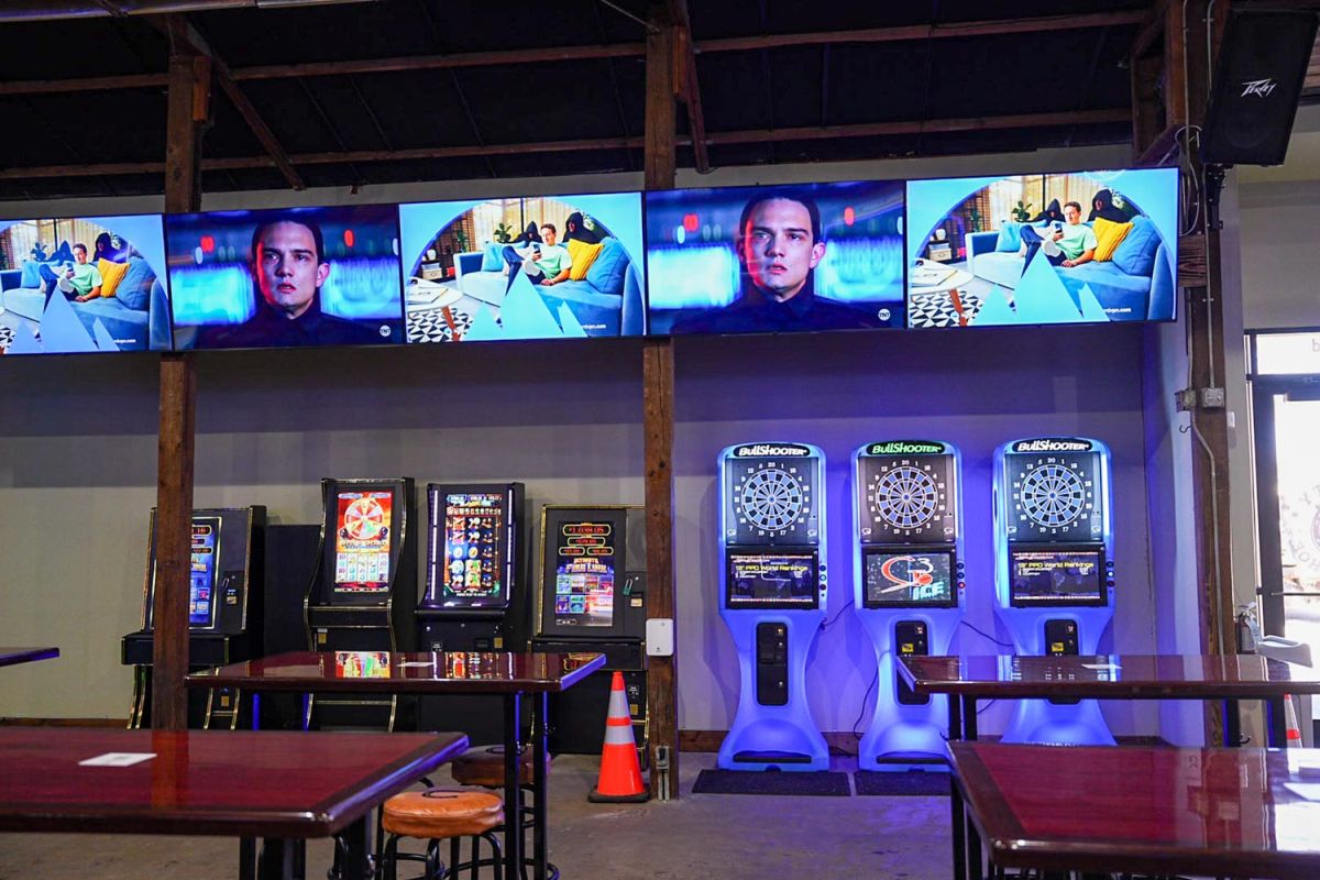 Wall-mounted screens above arcade machines and dartboards in a bar setting