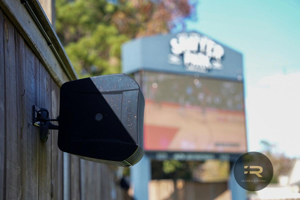 Outdoor speaker mounted on a wooden fence with a blurry sports screen in the background.