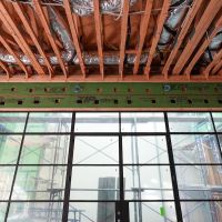 Framed ceiling joists above a large black-framed window wall on a construction site, exposed insulation ducts