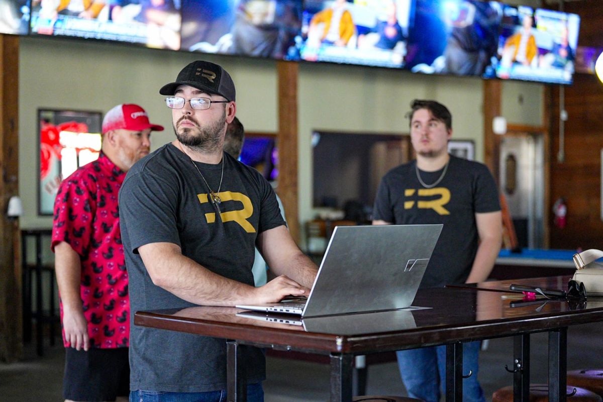 Team members wearing branded shirts gathered around a laptop in a sports bar