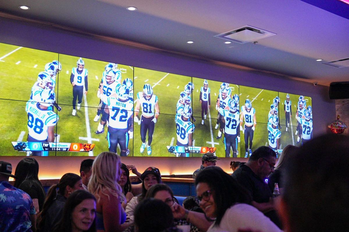 Wall of TVs showing a football team huddle in a crowded bar.