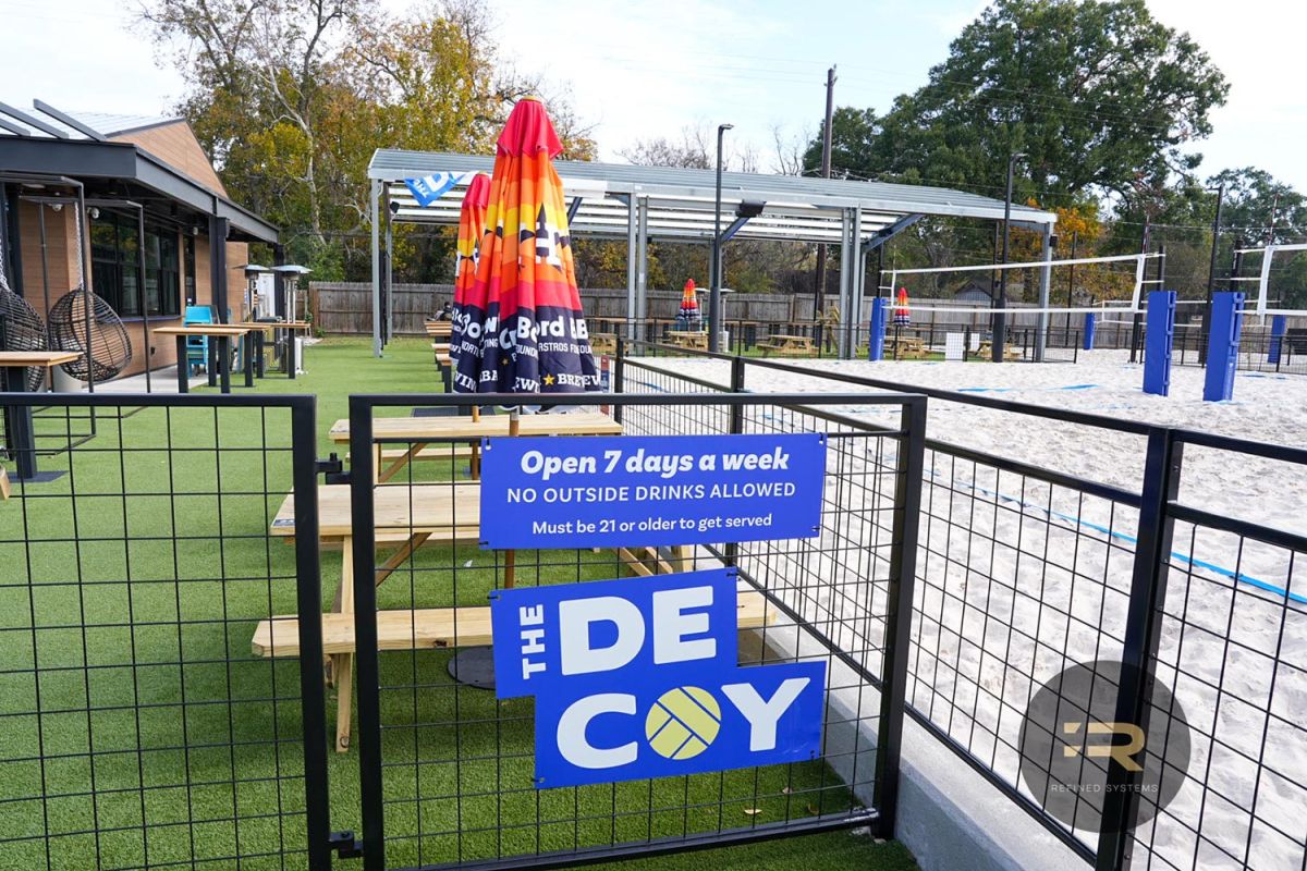 Outdoor entrance area with picnic tables, colorful umbrellas, and a sand volleyball court.