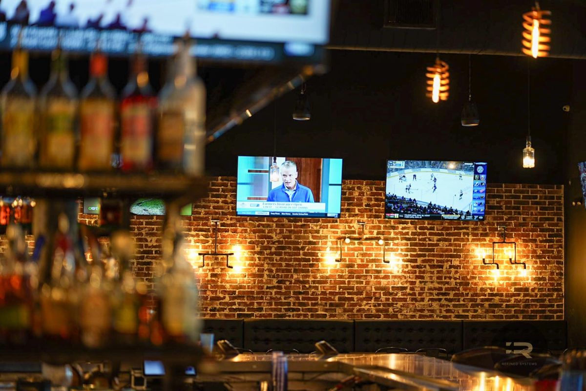 TVs showing a hockey game and interview in a bar with bottles and warm lighting.