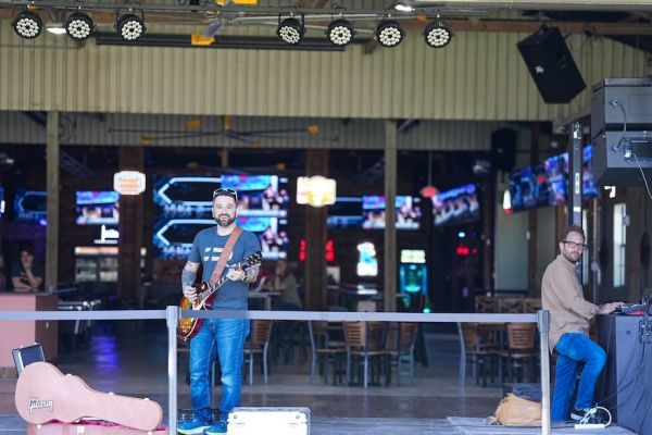 Musician with guitar performs on outdoor patio stage with professional lighting rig and screens visible in background.