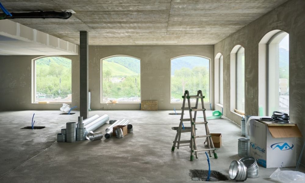 <span class="uk-h4">Interior of a building under construction with ductwork, ladder, and open windows showing green hills and blue sky outside.</span>