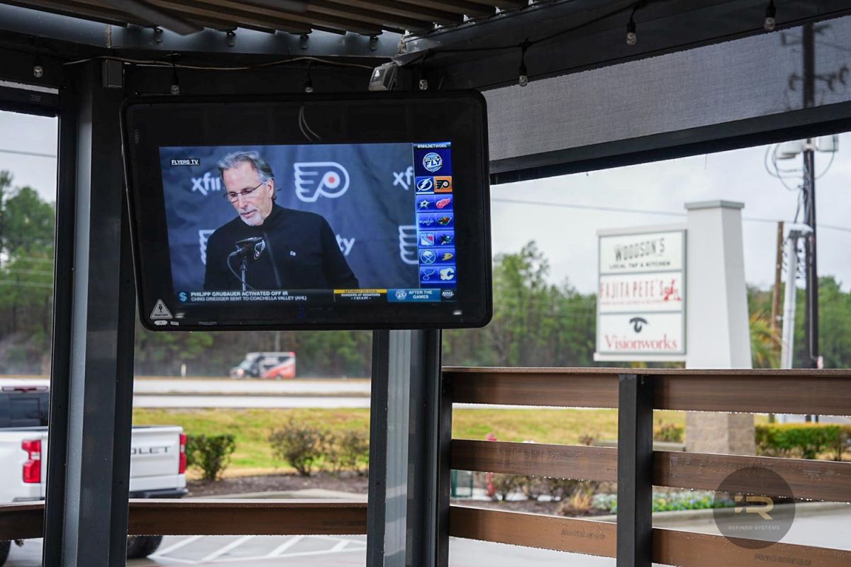Outdoor TV mounted under a covered patio showing a hockey interview.