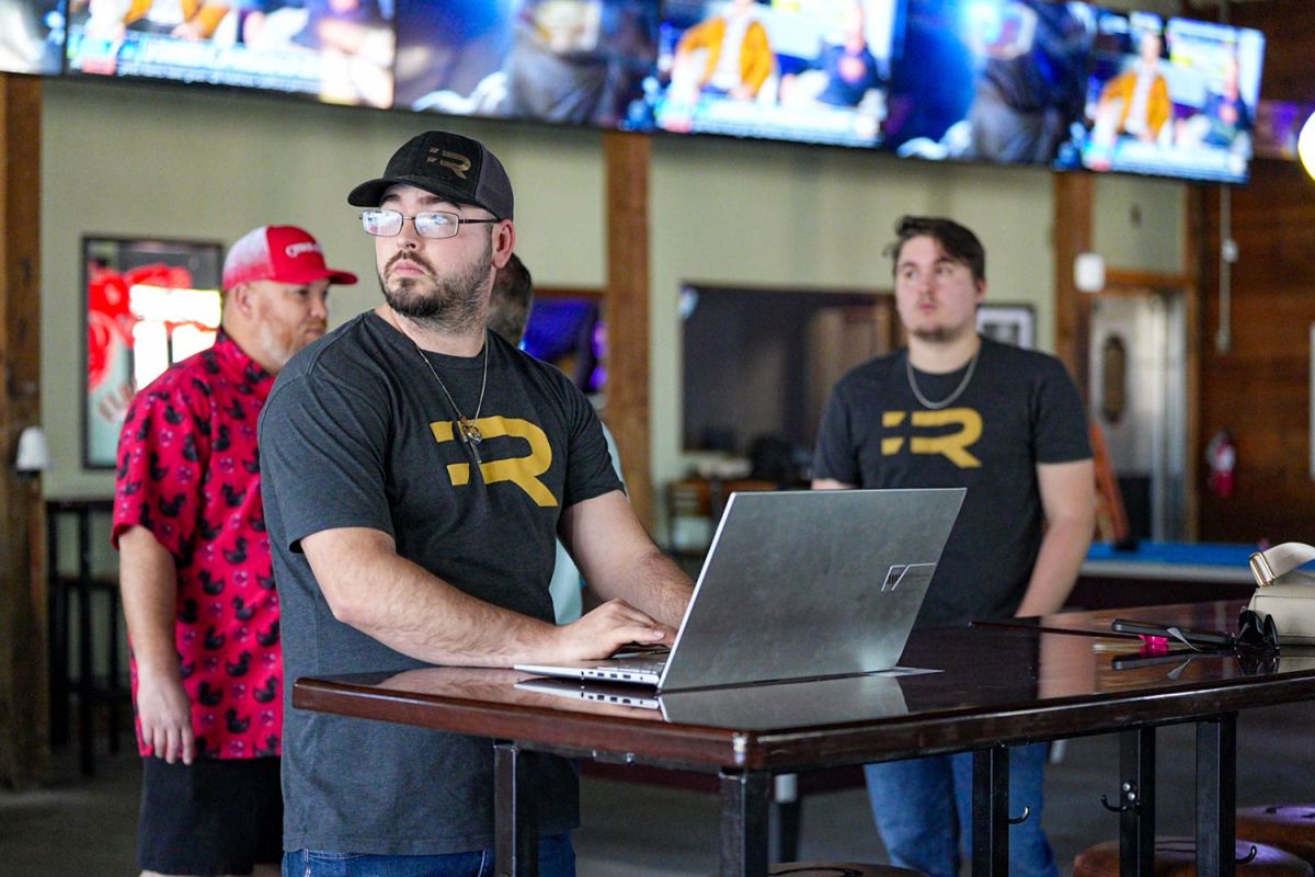 Team members wearing branded shirts gathered around a laptop in a sports bar