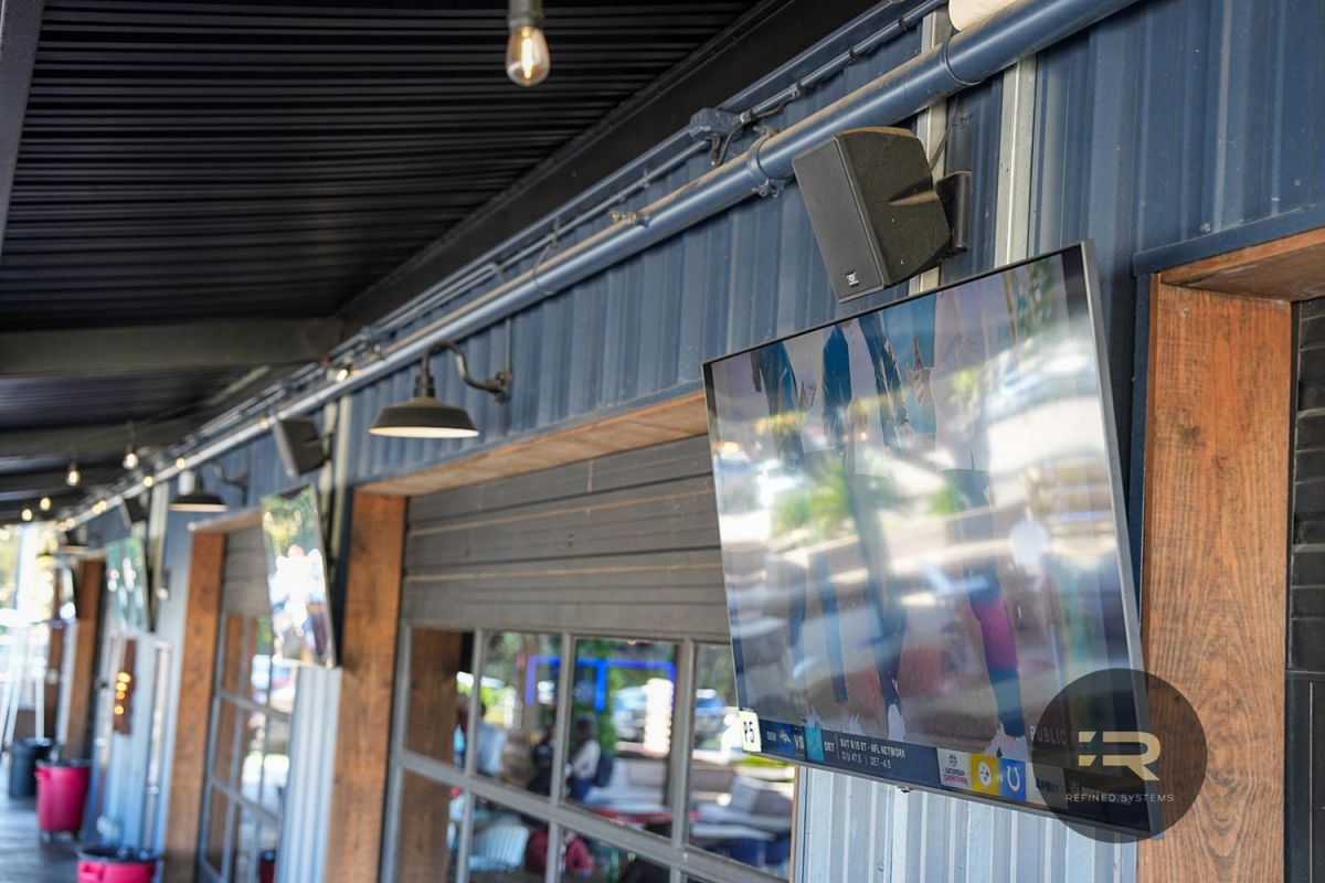 Multiple outdoor TVs mounted along a patio wall with black fixtures and speakers.