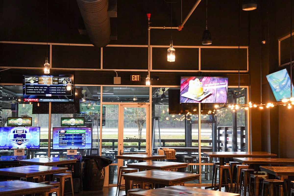 TVs and arcade machines near a glass exit with string lights and wooden tables.