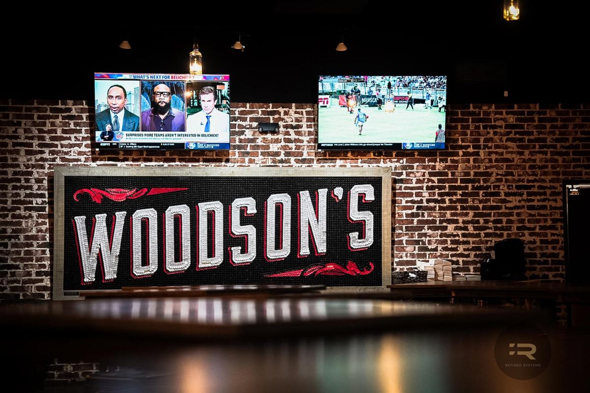Brick wall with two TVs showing sports and a large Woodson’s logo sign below.