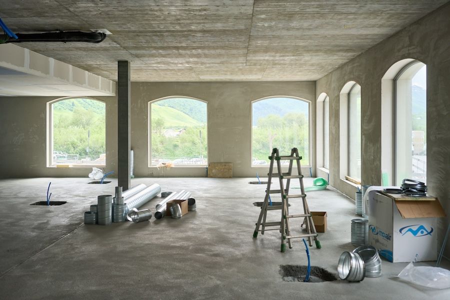 Interior of a building under construction with ductwork, ladder, and open windows showing green hills and blue sky outside.