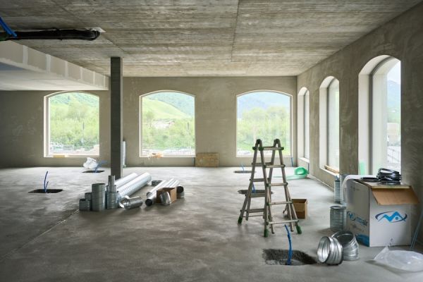 Interior of a building under construction with ductwork, ladder, and open windows showing green hills and blue sky outside.